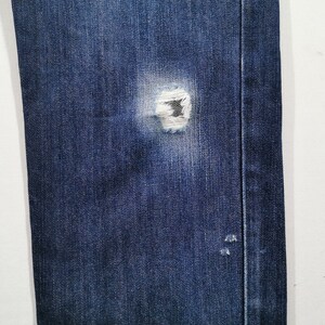 May include: Close-up of blue denim jeans with a distressed, ripped knee. The jeans show a worn appearance with visible fraying around the tear. The fabric has a classic, textured look.
