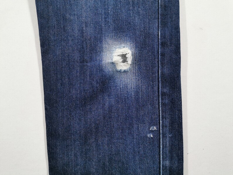 May include: Close-up of blue denim jeans with a distressed, ripped knee. The jeans show a worn appearance with visible fraying around the tear. The fabric has a classic, textured look.