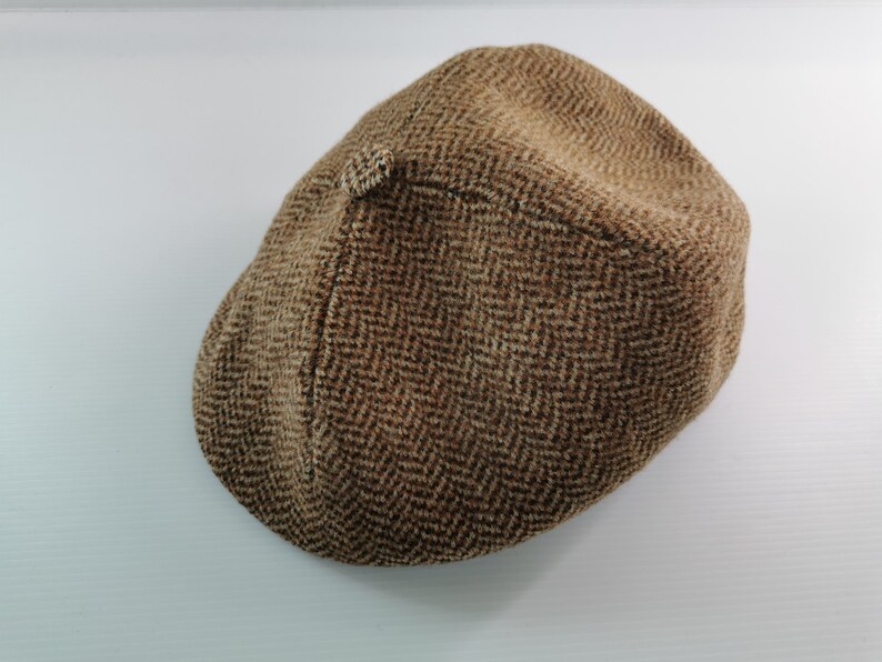 Op de afbeelding: Bruine tweed newsboy cap met een visgraatpatroon. De pet is gemaakt van een textuurstof en heeft een kleine, afgeronde klep. Een klassieke hoedstijl die geschikt is voor diverse gelegenheden.