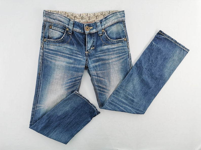 May include: Blue denim jeans with a faded wash and decorative stitching. The jeans feature a classic five-pocket design, a button and zipper closure, and a patterned waistband. The jeans are folded and laid flat, showcasing their details.