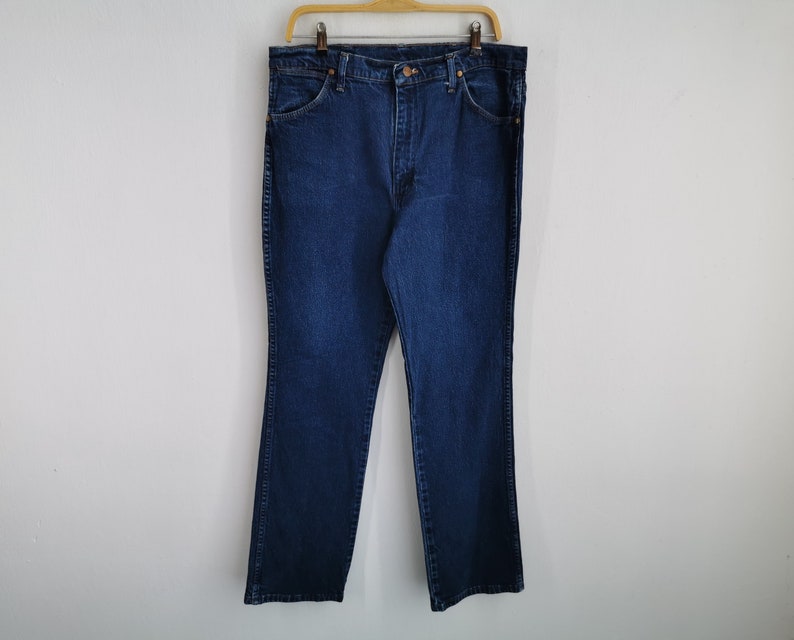 May include: Dark blue denim jeans hanging on a wooden hanger. The jeans have a classic five-pocket design, a button and zipper closure, and a straight leg fit. The jeans are made of a sturdy denim material and are suitable for everyday wear.