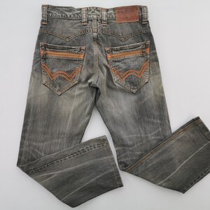 May include: Gray denim jeans with orange stitching details on the back pockets and a brown leather patch. The jeans have a faded, worn look and a classic five-pocket design. The back pockets have a unique, stylized design.
