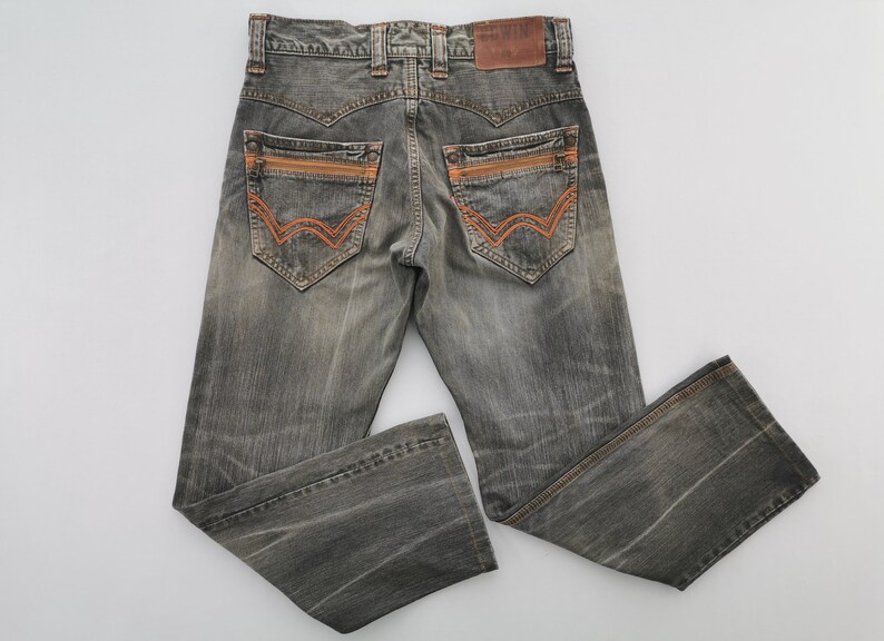 May include: Gray denim jeans with orange stitching details on the back pockets and a brown leather patch. The jeans have a faded, worn look and a classic five-pocket design. The back pockets have a unique, stylized design.