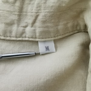 May include: A light beige shirt with a visible collar and a white fabric tag sewn into the neckline. The tag features the letter M in black print. A metallic tool with a textured grip is positioned horizontally, pointing toward the size tag.