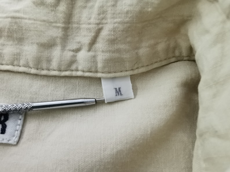 May include: A light beige shirt with a visible collar and a white fabric tag sewn into the neckline. The tag features the letter M in black print. A metallic tool with a textured grip is positioned horizontally, pointing toward the size tag.