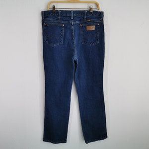 May include: Dark blue denim jeans hanging on a wooden hanger. The jeans have a classic five-pocket design, a brown leather patch with a logo, and copper-colored rivets. The Wrangler logo is visible on the back pocket.
