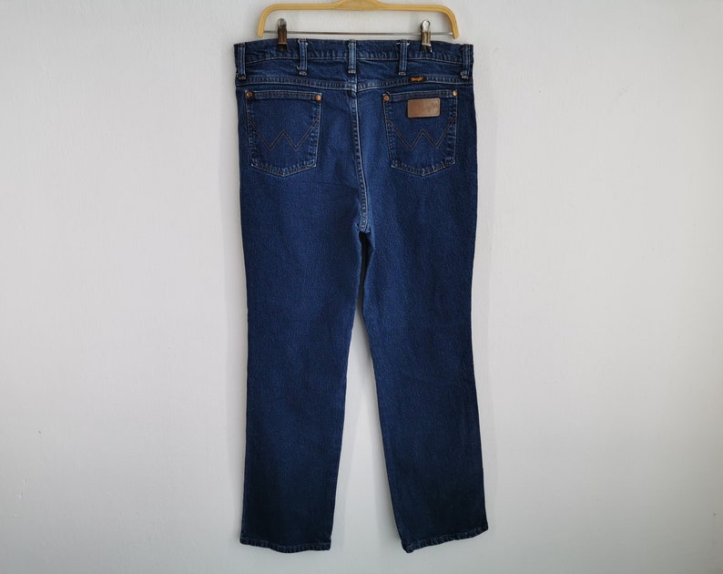 May include: Dark blue denim jeans hanging on a wooden hanger. The jeans have a classic five-pocket design, a brown leather patch with a logo, and copper-colored rivets. The Wrangler logo is visible on the back pocket.