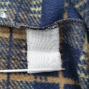Puede incluir: Primer plano de una secci&oacute;n de tela a cuadros azul, marr&oacute;n y beige. Una etiqueta blanca est&aacute; adherida a la tela. Una aguja plateada est&aacute; colocada cerca de la etiqueta.