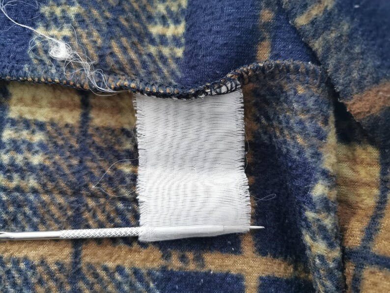 Puede incluir: Primer plano de una secci&oacute;n de tela a cuadros azul, marr&oacute;n y beige. Una etiqueta blanca est&aacute; adherida a la tela. Una aguja plateada est&aacute; colocada cerca de la etiqueta.