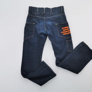 May include: Dark blue denim jeans with a straight leg design. The back right pocket features the orange text "STUDIO SEVEN CHILDHOOD DREAMS". The jeans have two back pockets and two side pockets. The waist has a button and belt loop closure.