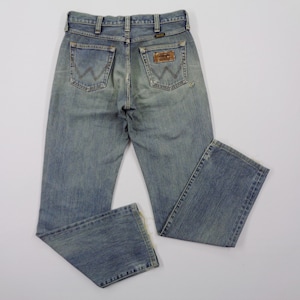 Pu&ograve; includere: Jeans di denim blu chiaro con un classico design a cinque tasche. I jeans presentano un lavaggio sbiadito e una toppa del marchio Wrangler sulla tasca posteriore. I jeans sono appoggiati su una superficie bianca.