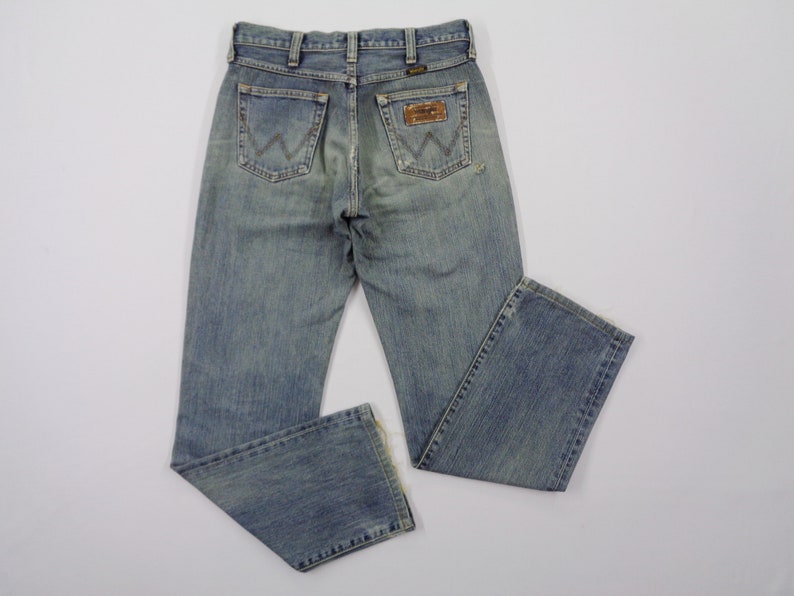 Pu&ograve; includere: Jeans di denim blu chiaro con un classico design a cinque tasche. I jeans presentano un lavaggio sbiadito e una toppa del marchio Wrangler sulla tasca posteriore. I jeans sono appoggiati su una superficie bianca.