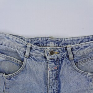 May include: Close-up of light blue acid wash denim jeans. The jeans have a button closure, belt loops, and front pockets. The denim has a faded, textured appearance.