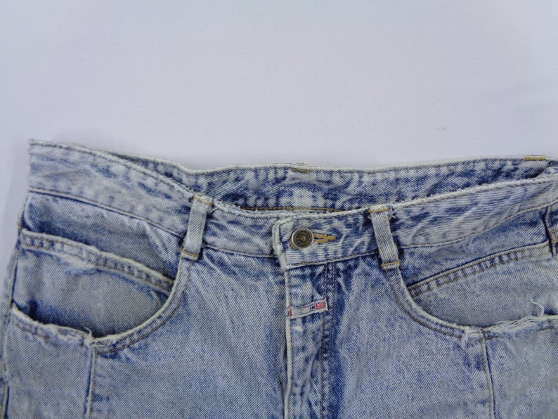May include: Close-up of light blue acid wash denim jeans. The jeans have a button closure, belt loops, and front pockets. The denim has a faded, textured appearance.