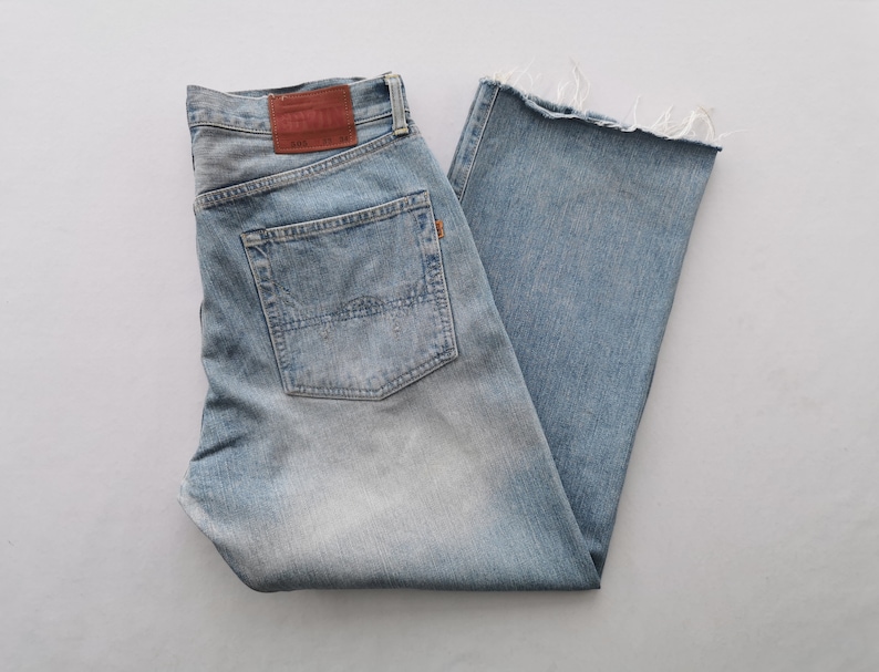 Puede incluir: Vaqueros de mezclilla azul claro con un lavado descolorido y un dobladillo sin rematar y deshilachado. Los vaqueros presentan un dise&ntilde;o cl&aacute;sico de cinco bolsillos, un parche de cuero marr&oacute;n y cierre de bot&oacute;n y cremallera. Los vaqueros est&aacute;n doblados cuidadosamente.