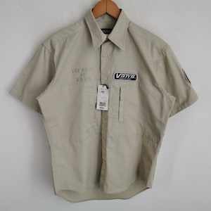 May include: Tan short-sleeve button-up shirt with a collar, hanging on a wooden hanger. The shirt features the text "THE SOUL OF VANS" and a Vans logo patch. A price tag hangs from the shirt.
