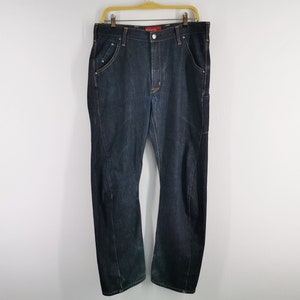 May include: Dark wash denim jeans hanging on a wooden hanger. The jeans have a classic five-pocket design with a straight leg and a slight bootcut. The stitching is a contrasting gold colour.