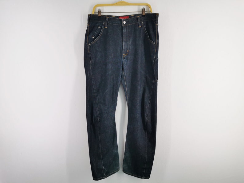 May include: Dark wash denim jeans hanging on a wooden hanger. The jeans have a classic five-pocket design with a straight leg and a slight bootcut. The stitching is a contrasting gold colour.