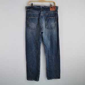 May include: Blue denim jeans with a faded wash, hanging on a wooden hanger. The jeans have a classic five-pocket design, a button and zipper closure, and a leather patch on the back. The jeans are a medium wash with some fading and distressing.