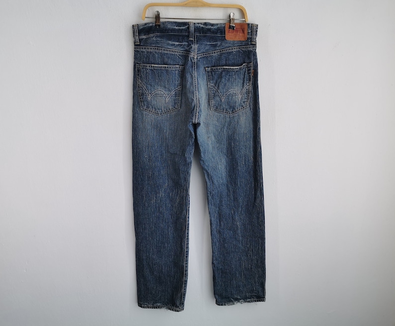 May include: Blue denim jeans with a faded wash, hanging on a wooden hanger. The jeans have a classic five-pocket design, a button and zipper closure, and a leather patch on the back. The jeans are a medium wash with some fading and distressing.