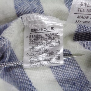 Może przedstawiać: Biało-niebieska odzież w paski z białą metką z instrukcją prania w języku japońskim oraz drugą metką z napisem Made in China. Metalowe narzędzie o teksturowanej rączce wskazuje na japoński tekst znajdujący się na metce pielęgnacyjnej.