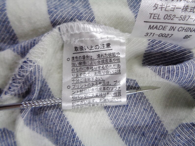 Może przedstawiać: Biało-niebieska odzież w paski z białą metką z instrukcją prania w języku japońskim oraz drugą metką z napisem Made in China. Metalowe narzędzie o teksturowanej rączce wskazuje na japoński tekst znajdujący się na metce pielęgnacyjnej.