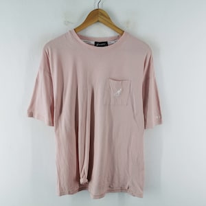 May include: A light pink t-shirt with a pocket on the chest featuring a white kangaroo logo. The shirt has short sleeves and a crew neck. The brand name is visible on the collar. The shirt is hanging on a wooden hanger.