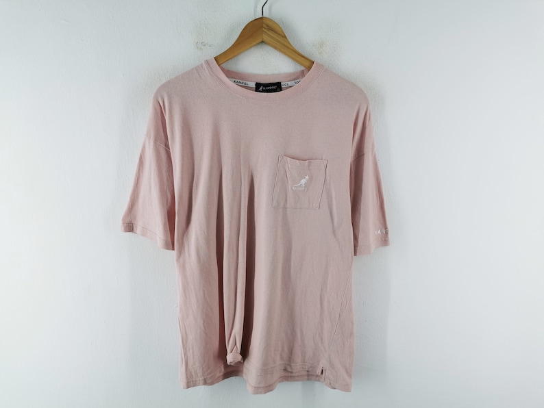 May include: A light pink t-shirt with a pocket on the chest featuring a white kangaroo logo. The shirt has short sleeves and a crew neck. The brand name is visible on the collar. The shirt is hanging on a wooden hanger.