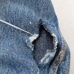 May include: Close-up of distressed blue denim fabric with a tear. A silver tool is inserted into the tear, highlighting the frayed edges and the fabric's texture. The denim shows signs of wear and tear, with visible stitching.