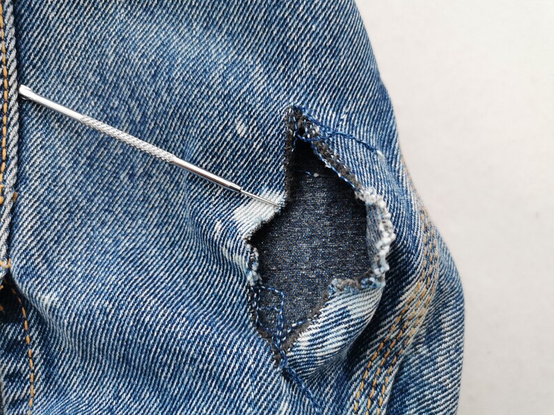 May include: Close-up of distressed blue denim fabric with a tear. A silver tool is inserted into the tear, highlighting the frayed edges and the fabric's texture. The denim shows signs of wear and tear, with visible stitching.