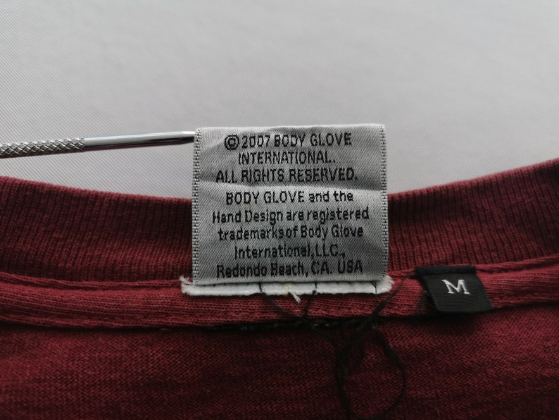 May include: A close-up of a burgundy t-shirt label with the text "&copy;2007 BODY GLOVE INTERNATIONAL. ALL RIGHTS RESERVED." The label also includes the Body Glove trademark and the location "Redondo Beach, CA. USA."