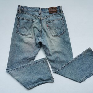 May include: Light blue denim jeans with a faded wash. The jeans have a classic five-pocket design, a brown leather patch, and a red tag on the back pocket. The jeans have a bootcut leg opening.