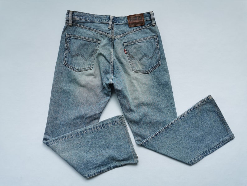 May include: Light blue denim jeans with a faded wash. The jeans have a classic five-pocket design, a brown leather patch, and a red tag on the back pocket. The jeans have a bootcut leg opening.