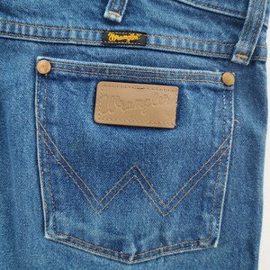 May include: Blue denim jeans featuring a back pocket with a tan leather patch embossed with the Wrangler logo. The pocket includes copper-colored rivets at the top corners and a signature W-shaped stitch pattern in gold thread throughout the fabric.