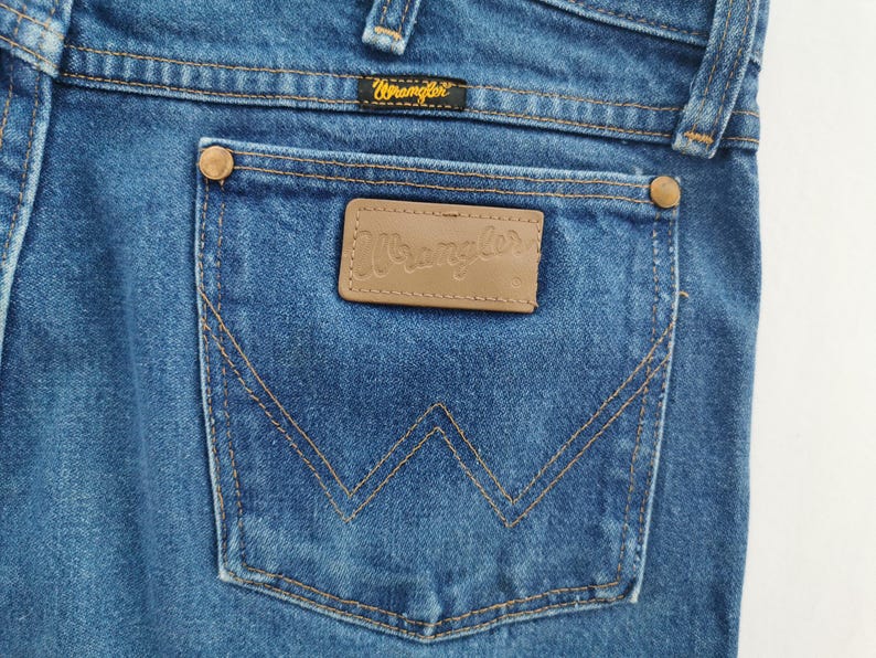 May include: Blue denim jeans featuring a back pocket with a tan leather patch embossed with the Wrangler logo. The pocket includes copper-colored rivets at the top corners and a signature W-shaped stitch pattern in gold thread throughout the fabric.