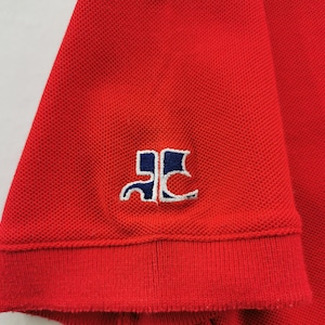 K&ouml;nnte beinhalten: Nahaufnahme eines roten Polo-Shirt-&Auml;rmels. Der &Auml;rmel hat einen gerippten Bund und ein kleines gesticktes Logo in Wei&szlig; und Marineblau. Der Stoff hat eine strukturierte Piqu&eacute;-Webart.