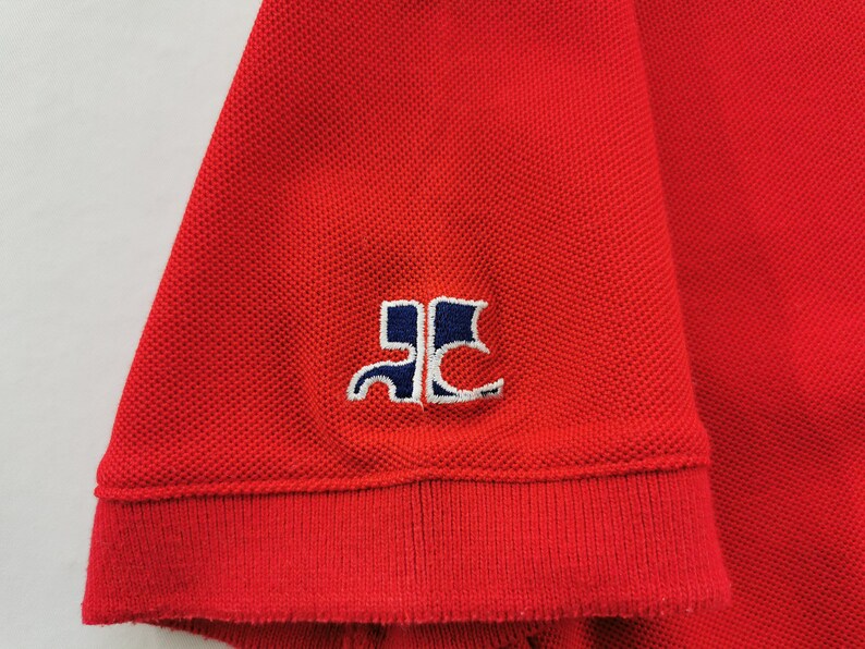 K&ouml;nnte beinhalten: Nahaufnahme eines roten Polo-Shirt-&Auml;rmels. Der &Auml;rmel hat einen gerippten Bund und ein kleines gesticktes Logo in Wei&szlig; und Marineblau. Der Stoff hat eine strukturierte Piqu&eacute;-Webart.