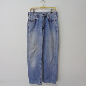 May include: Light blue denim jeans with a faded wash and a distressed knee detail. The jeans are hanging on a yellow hanger against a plain white background. The jeans have a classic five-pocket design.