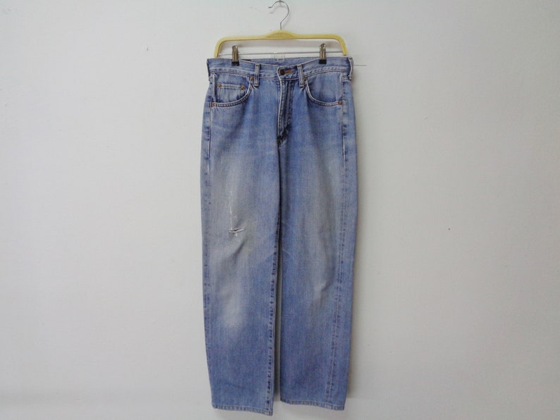 May include: Light blue denim jeans with a faded wash and a distressed knee detail. The jeans are hanging on a yellow hanger against a plain white background. The jeans have a classic five-pocket design.