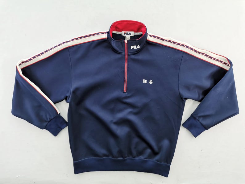 Pu&ograve; includere: Giacca da tuta FILA blu navy con un design a righe rosse e bianche sulle maniche e sul colletto. La giacca ha una chiusura a mezza zip e il logo FILA sul colletto. Caratteri giapponesi visibili sul petto sinistro.