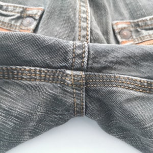May include: Close-up of gray denim jeans with orange stitching and accents. The jeans have a classic five-pocket design with metal rivets and a button closure. The stitching is visible on the seams and pockets.