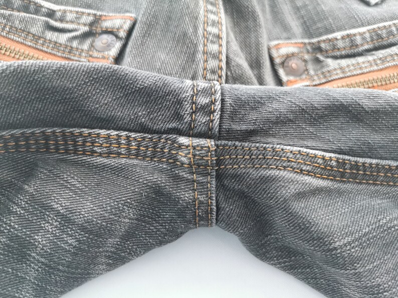 May include: Close-up of gray denim jeans with orange stitching and accents. The jeans have a classic five-pocket design with metal rivets and a button closure. The stitching is visible on the seams and pockets.