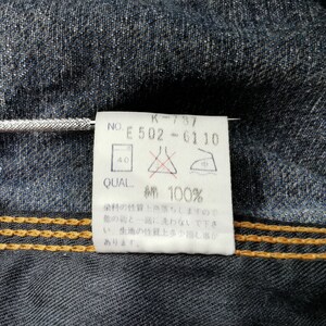 May include: Close-up of a denim garment with a white tag attached. The tag displays text including "K-797", "E 502-6110", and "綿 100%", indicating the fabric composition. The denim has visible stitching and a textured appearance.