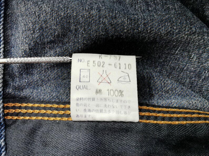 May include: Close-up of a denim garment with a white tag attached. The tag displays text including "K-797", "E 502-6110", and "綿 100%", indicating the fabric composition. The denim has visible stitching and a textured appearance.