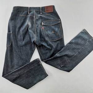 May include: Dark wash denim jeans with a classic five-pocket design, featuring a unique pocket on the left leg. The jeans have a brown leather patch and a red tag on the back pocket. The jeans are laid flat on a white surface.
