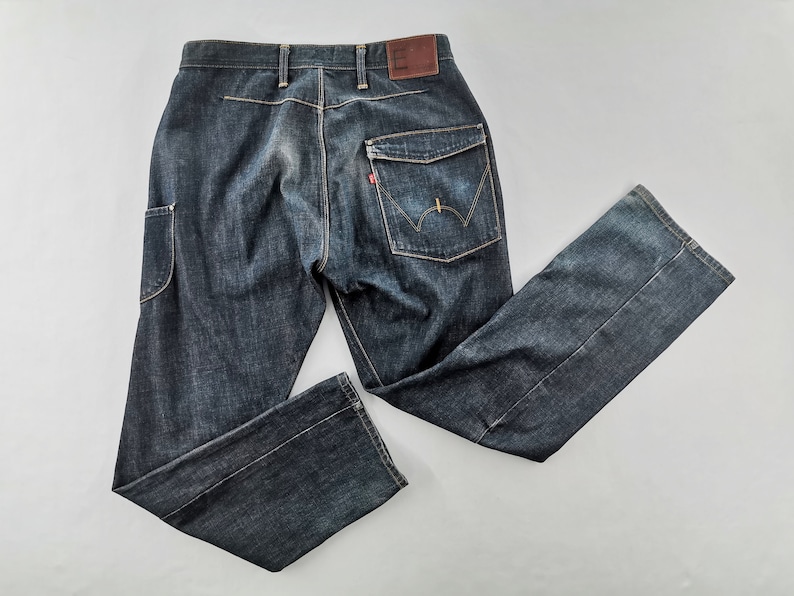 May include: Dark wash denim jeans with a classic five-pocket design, featuring a unique pocket on the left leg. The jeans have a brown leather patch and a red tag on the back pocket. The jeans are laid flat on a white surface.