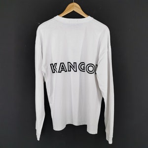 Op de afbeelding: Wit shirt met lange mouwen met het woord "KANGOL" in zwart op de rug gedrukt. Het shirt hangt aan een houten hanger tegen een donkere achtergrond. De mouwen zijn lang en de stof lijkt licht van gewicht.