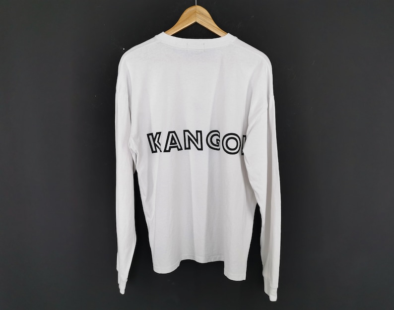 Op de afbeelding: Wit shirt met lange mouwen met het woord "KANGOL" in zwart op de rug gedrukt. Het shirt hangt aan een houten hanger tegen een donkere achtergrond. De mouwen zijn lang en de stof lijkt licht van gewicht.