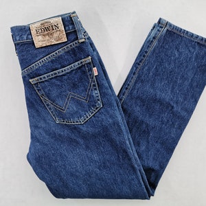 Puede incluir: Vaqueros de mezclilla azul oscuro con un dise&ntilde;o cl&aacute;sico de cinco bolsillos. Cuenta con un parche de cuero marr&oacute;n con la marca "EDWIN" en la parte trasera y una etiqueta roja. Los vaqueros tienen un corte recto y est&aacute;n doblados.