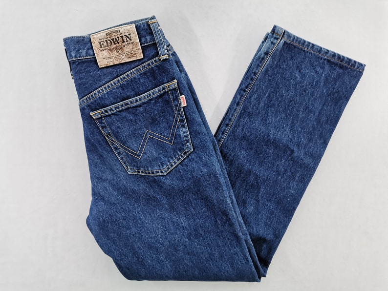 Puede incluir: Vaqueros de mezclilla azul oscuro con un dise&ntilde;o cl&aacute;sico de cinco bolsillos. Cuenta con un parche de cuero marr&oacute;n con la marca "EDWIN" en la parte trasera y una etiqueta roja. Los vaqueros tienen un corte recto y est&aacute;n doblados.